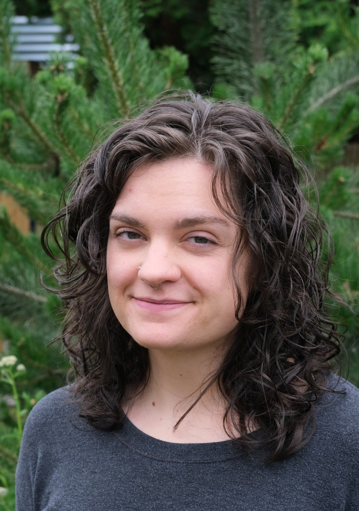Woman smiling in a friendly manner in front of an evergreen tree.
